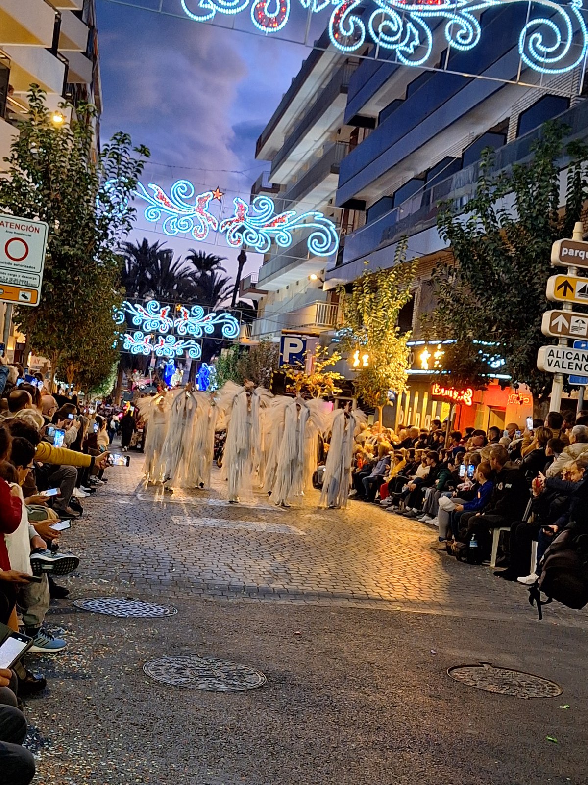 Dancers in white costumes under the Christmas lights on the Three Kings parade route, crowds either side